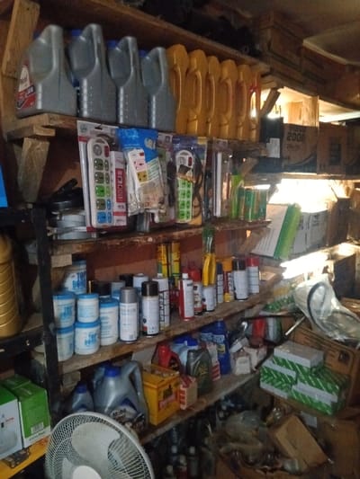 This image depicts an interior view of a cluttered storage or retail space, likely in Akodo, Nigeria, characterized by multiple wooden shelves heavily laden with various items.

The top shelf contains rows of plastic containers, including several grey bottles with "Mobil 5W-40" branding, and numerous yellow, unbranded plastic containers, consistent with lubricants or automotive fluids.

The second shelf displays an assortment of packaged goods, primarily consumer electronics or hardware such as multiple power strips, remote controls in blister packaging, and other small items, along with green aerosol cans.

The third shelf holds containers labeled "TOP BOND," several white, blue, and black cylindrical containers, and numerous aerosol cans of various colors (red, white, black, grey), suggesting spray paints, adhesives, or sealants.

The fourth shelf, partially visible, contains additional grey plastic bottles resembling motor oil, a yellow rectangular box, red and blue plastic bottles, and stacks of green-and-white boxed items, possibly automotive parts or filters.

The lower foreground shows a white electric stand fan. The surrounding environment includes various cardboard boxes, plastic bags, and miscellaneous items scattered on the floor and lower shelves, indicating a densely packed storage area. Illumination is provided by artificial light sources, creating bright reflections and some shadowed areas within the scene. No individuals are present.