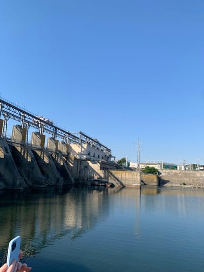A wide shot captures a hydroelectric dam on a clear, sunny day. The dam, constructed of concrete and steel, spans across a body of water. The water's surface reflects the dam's structure and the bright blue sky. In the foreground, a person's hands hold a light blue smartphone, angled towards the dam. The dam itself is a massive structure with several gates and machinery visible on top. The sky is a cloudless expanse of blue, with a few distant birds visible. Beyond the dam, a few buildings and power lines are present, suggesting a surrounding industrial or urban area. The image appears to depict the dam in Ustia, Moldova, a significant infrastructure project.