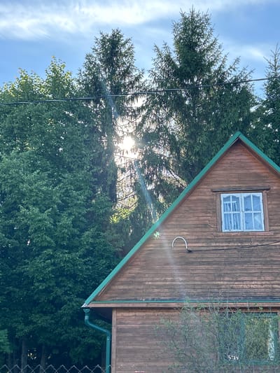 The image depicts a wooden house with a green roof in the village of Московский Литератор Снт, Russia. The sun is shining brightly through tall evergreen trees, creating a lens flare effect. A window with white curtains is visible on the upper floor of the house, and a white pipe is attached to the exterior wall. A green gutter system is installed along the roofline. In the foreground, a chain-link fence and some bushes are partially visible. The sky is blue with scattered clouds.