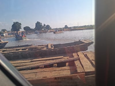 This image captures a busy waterfront scene in Nemnei, Nigeria, under a clear, light blue sky. In the immediate foreground, a weathered wooden pier or dock, constructed from rough planks, extends across the lower portion of the frame. The bow section of a dark, old wooden boat is partially visible alongside this structure. The view is partially obstructed by a dark, diagonal element in the lower left and a vertical dark object along the far right edge, suggesting the photograph was taken from within an enclosure.
The central area of the image is dominated by a body of water, likely a river, upon which multiple wooden boats are active. A prominent green motorboat, visibly overloaded with approximately 15-20 individuals, is moving from left to right, leaving a distinct white wake in its path. Other wooden boats are distributed across the water, some appearing stationary near the distant bank.
The far bank features a dirt shoreline, several large green trees, and a collection of parked vehicles, including white vans, indicating a land-based connection point. A faint communication tower is visible in the far background against the clear sky. The scene depicts a functional water transport hub.
