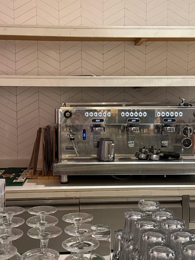 The image depicts the interior of a cafe, likely in Ras Al-Khaimah, United Arab Emirates, focusing on a professional espresso machine and glassware. The espresso machine, a prominent chrome appliance, is situated on a counter, displaying multiple brewing heads and a control panel with digital readouts and illuminated buttons. To the left of the machine, a metal milk pitcher and portafilters are visible on the counter. Behind the machine, a tiled backsplash with a chevron pattern lines the wall. Two shelves, one wooden and one marble, are mounted on the wall above the machine. In the foreground, stacks of clear glassware, likely for serving drinks, are arranged on a counter. The overall impression is one of a clean and functional cafe environment. There are no people visible in the image.