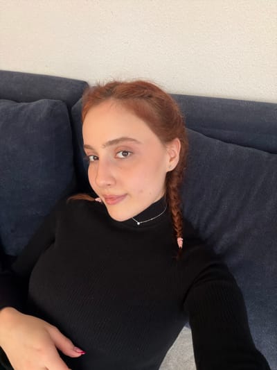 A young female, appearing to be in an indoor setting, is seated or reclining on a dark blue fabric sofa. Her reddish-brown hair is styled in two braids, secured with pink ties, with one braid visible resting on her right shoulder. She wears a black ribbed turtleneck sweater and a thin silver necklace featuring a small, square-shaped pendant. A small silver stud earring is visible in her right ear. Her left hand, with fingernails painted pink in a French tip style, rests on her lap or abdomen. She looks directly at the camera with a slight smile. The background consists of the sofa and a plain, light-colored wall. There are no visual cues within the image to specify a location in the city of Eschau, France, or any other geographical detail.