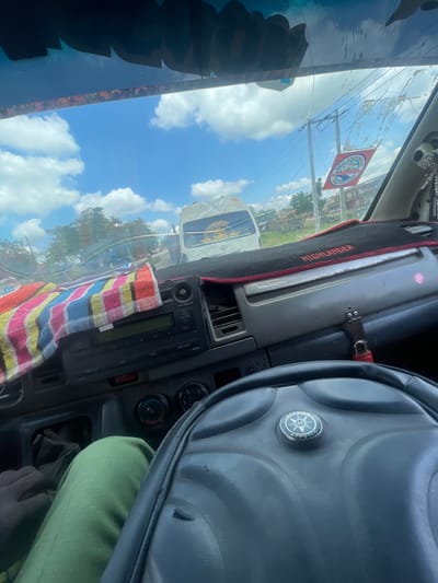 The image is taken from the interior of a vehicle, looking forward through the windshield. The dashboard is visible, with a colorful striped cloth draped over the center console, partially obscuring a car stereo. The steering wheel is in the foreground, to the right. Outside the vehicle, a bright blue sky with scattered white clouds is visible. Several vehicles, including a white bus with signage, are seen on a road, suggesting a city or town environment. Trees and buildings are present in the background. A red and white circular sticker with text is attached to the windshield. The lighting suggests it is daytime. Based on the context provided, the location is the city of Ikire, Nigeria.