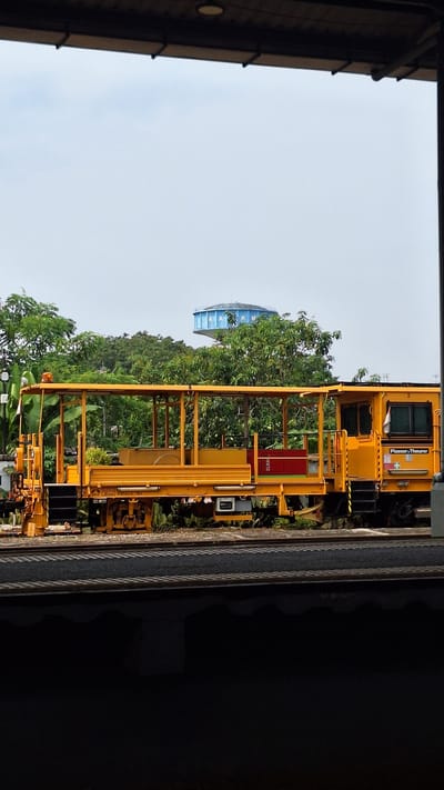 The image shows a yellow maintenance vehicle on a railway track, framed by the structure of a building, possibly a warehouse or industrial shed, with the sky visible in the distance. The vehicle has a complex frame, and is parked on tracks with green vegetation visible nearby. In the background, beyond the railway and vegetation, a water tower with "KARAWANG" inscribed is noticeable. The scene is likely in or around Karawang, Indonesia, due to the prominence of the city's name on the water tower.