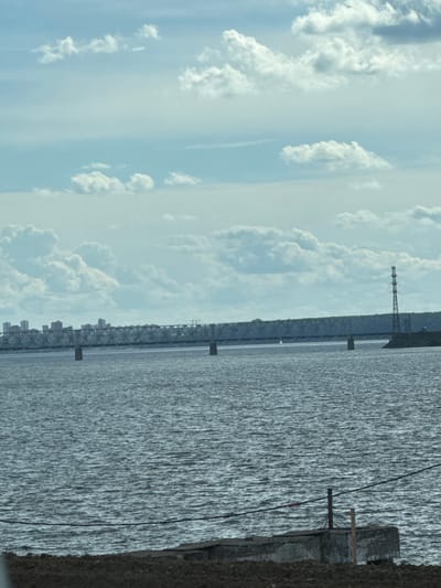 The image depicts a wide body of water, likely a river, with a long metal truss bridge spanning across it. In the distance, a city skyline with several tall buildings is visible on the far bank, suggesting an urban or suburban setting. A tall, lattice-like power transmission tower stands on the right bank, closer to the foreground, with cables extending from it. The water is choppy, indicating a slight breeze, and the sky is partly cloudy with soft, white clouds against a light blue background. The foreground shows a rough, earthy bank with some concrete structures and what appears to be dark soil or gravel. The overall impression is a landscape view of a bridge and city across a body of water, characteristic of a riverside urban area. The location is specified as Palniky, Russia.