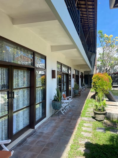 The image depicts an outdoor hallway or walkway adjacent to a building with several rooms. The building has a white exterior and large glass doors with dark brown frames. The doors are adorned with patterned curtains featuring leaves and branches. The walkway is paved with square, brown tiles and is lined with potted plants, including a tall snake plant in a teal pot. Several white chairs are placed along the walkway.
Further down the walkway, there are more white chairs and a large, lush green shrub with reddish tips. To the right of the walkway, a body of water, possibly a swimming pool, is visible, along with lounge chairs and some greenery. A wooden-beamed roof structure extends over the walkway, supported by white columns. The sky is clear and blue, and sunlight casts shadows on the ground.
In the background, a tree with yellow flowers is visible. The scene suggests a resort, guesthouse, or a similar type of accommodation. The location is identified as Denpasar Barat, Indonesia. There are no people visible in the image, and no actions or interactions are occurring. The environment is tranquil and well-maintained.