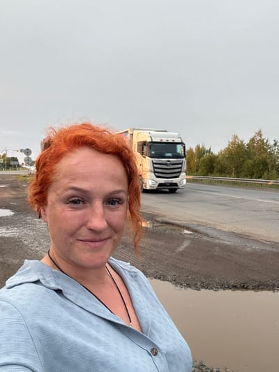 This media file captures a close-up portrait of a woman with bright red, curly hair. She is wearing a light blue button-up shirt, and a thin black cord necklace is visible. Her expression is neutral. In the background, a white truck is parked on a wet road, suggesting recent rain. Trees and a road barrier are visible further behind the truck. The scene appears to be an outdoor setting, possibly a roadside in Sovkhoznyi, Russia, given the provided location context. The lighting indicates it may be daytime, with overcast or diffused light. The wet road and puddles suggest damp or rainy weather. No visible text is present in the image.