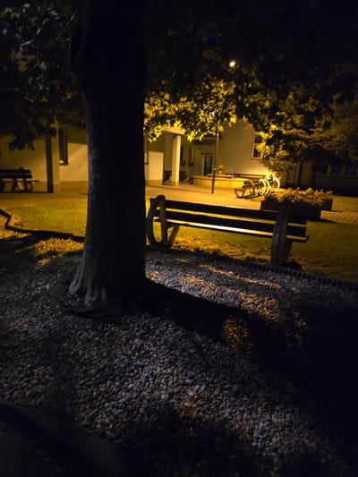 In Kostelec na Hané, Czechia, at night, a tree trunk dominates the foreground, casting a shadow across a gravel path and a grassy area. To the right of the tree, a wooden bench with large wagon wheel-like supports is positioned on the grass. Further back, a planter box filled with flowers sits near another bench. Behind the benches and planter, a building with windows is visible, illuminated by artificial light. Several bicycles are parked in front of the building. The overall scene suggests an outdoor public space, likely a park or courtyard, during nighttime.