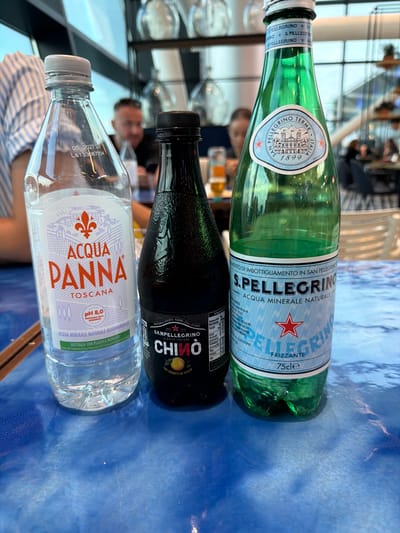 This is a vertical, eye-level medium shot of three bottles of water and a bottled beverage on a blue, textured surface, likely a table. In the foreground, a clear plastic bottle of Aqua Panna Toscana is visible, with its cap and label clearly showing the brand name and "Toscana" in orange lettering, along with a crest. Behind it, a dark green glass bottle with a black cap, labeled Sanpellegrino Chino, is tilted. The label prominently features the Sanpellegrino logo and "Chino" in a yellow circle with a lemon graphic. The farthest bottle, also green glass and topped with a white cap, is labeled S.Pellegrino with a circular emblem that says "San Pellegrino Terme Italia 1899". The background is blurred, showing hints of people, a bright interior space, and possibly a window with some vertical elements. The overall impression is that of a beverage selection at a restaurant or bar. The lighting suggests daytime. Visible text includes brand names, product details, and nutritional information.