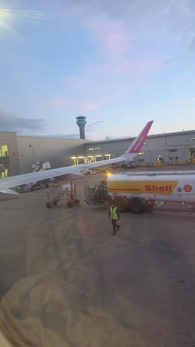 The image, oriented as a landscape view, captures an airport scene from inside an airplane, looking out a window onto the tarmac. A significant portion of the airplane's wing extends from the lower-right to the upper-left, partially obscuring the view below. The aircraft's tail, visible on the left side, is magenta with the white text "wizzair.com" vertically printed on it.
Adjacent to the airplane, a large yellow and white Shell fuel tanker truck is parked, suggesting a refueling operation. The word "Shell" is prominently displayed on the side of the truck in red lettering, along with safety warnings like "FLAMMABLE" and "NO SMOKING". A ground crew member, wearing a bright yellow or green high-visibility vest, stands on the tarmac near the truck and the aircraft wing.
In the background, a distinctive air traffic control tower with a dark, possibly glass, cab is visible. Behind the aircraft and truck, a modern airport terminal building with multiple windows and roof structures can be seen. The tarmac is dark gray with visible lines and markings.
The sky, visible in the upper-right portion, shows soft gradients of light blue and pink, indicating either dawn or dusk. The overall lighting is dim but clear, suggesting favorable weather conditions. The scene depicts standard ground operations at an airport, specifically refueling, likely at Luton, United Kingdom.