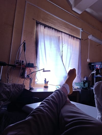 The image depicts an indoor scene, possibly a workshop or a study, with a person's legs and feet prominently in the foreground, resting on a white surface. The individual is wearing light-colored pants and yellow socks. The environment appears somewhat cluttered with various objects, including a desk with pens in a holder, a lamp, electrical outlets, and what seems to be a drill press in the background. A window with sheer curtains is visible, letting in natural light. The walls are textured and painted in a warm, muted tone. There is no specific indication of the city of Kobryn, Belarus, or any other identifiable geographical markers within the frame.