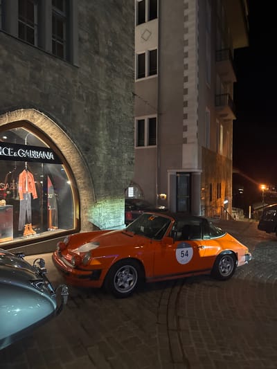 The image displays a nighttime street scene in Sankt Moritz, Switzerland. The central focus is an orange Porsche 911 Targa, identifiable by its black removable roof section and "Targa" script on the rear window. This vehicle features a white circular race number "54" on its driver's side door and is equipped with multi-spoke alloy wheels. It is parked on a cobblestone street.

In the immediate foreground, partially visible on the left, is the front bumper and a portion of the hood of a silver-grey classic car, also parked on the cobblestones.

To the left of the orange Porsche, a stone-faced building occupies the frame. Its lower section features a large, arched display window illuminated from within. The window prominently displays the "Dolce & Gabbana" logo. Inside, an orange jacket, dark trousers, and brown boots are arranged on mannequins. Above the storefront, the stone facade continues, featuring a dark, rectangular window on an upper floor.

To the right of the orange Porsche, a lighter-colored building stands with distinct decorative quoining on its corner. This building has multiple dark, rectangular windows and balconies on its right elevation. An entrance is recessed into its lower facade. Beyond this building, a cobblestone street slopes downward, and a streetlight is visible in the distance on the right. A dark-colored vehicle is partially visible in the far right background. Reflections of additional vehicles, including one with the number "11", are discernible in the storefront glass. No individuals are visible in the scene.