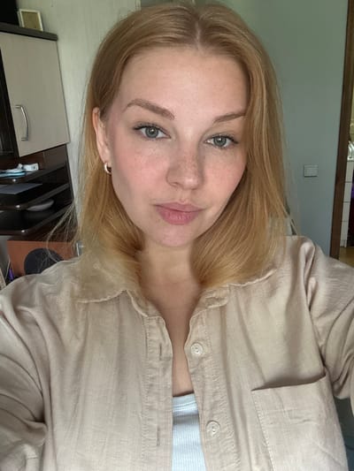 This is a waist-up, selfie-style photograph of a young woman with blonde hair and fair skin, wearing a beige button-up shirt over a white tank top. She has light blue eyes and freckles across her nose and cheeks. She is wearing small hoop earrings. The background appears to be an indoor domestic setting, possibly a bedroom or living area. To the left, a portion of a wooden cabinet with a silver handle is visible. To the right, a doorframe and a light switch on the wall are discernible. The lighting suggests it is daytime. The image does not contain any specific details that would place it in Yaroslavl, Russia, or any other particular city.