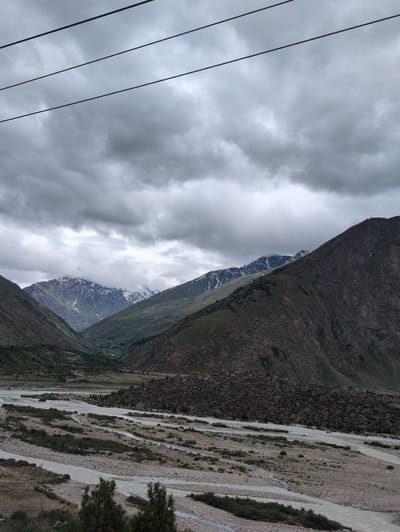 A panoramic view captures the rugged, mountainous terrain surrounding the town of Darcha, India, under a dramatic sky filled with heavy, dark clouds. In the foreground, a wide, braided riverbed dominates the landscape, with multiple channels of murky water flowing between islands of gravel and sparse, low-lying vegetation. The riverbanks are lined with rocks and some scrubby bushes.
The middle ground transitions into steep, rocky slopes covered with patches of green vegetation. These slopes rise to meet the imposing mountains that form the backdrop. The mountains are a mix of dark rock and scree, with some peaks showing caps of snow, even under the overcast conditions. A valley stretches between the mountains, gradually receding into the distance.
In the far distance, a few scattered structures, possibly houses or buildings, are visible nestled within the valley, indicating the presence of human habitation. Overhead, four distinct, dark power lines stretch across the frame, creating a linear element that contrasts with the natural curves of the landscape. The overall impression is one of a vast, untamed, and somewhat somber natural environment.