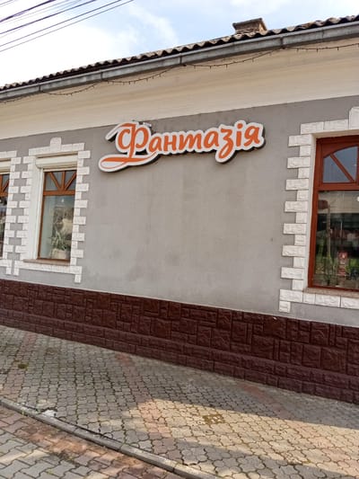 The image shows the exterior of a building with a sign that reads "Фантазія" (Fantaziya) in an orange, stylized font with a white outline. The building has a gray textured wall with decorative white brick accents around two windows and a brown tiled base. The window on the left is covered by a white roller shutter, and the one on the right has a wooden frame and is reflecting the surroundings. Above the windows, there's a white cornice. The roof is visible at the top, with some wires running across the sky, which is partly cloudy. The ground in front of the building is paved with interlocking concrete blocks. The scene appears to be outdoors during the daytime. There are no people visible. The overall impression is of a commercial establishment, possibly a shop or a business, in Tiachiv, Ukraine.