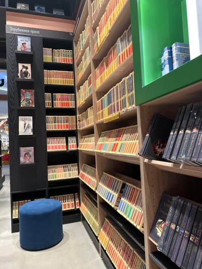 The image shows a section of a bookstore in Mukhanovo, Russia, featuring rows of books on shelves. On the left, a black bookshelf displays several books vertically, including titles with Russian text like "Зарубежная проза" (Foreign Prose). A blue ottoman sits in the foreground. To the right, wooden shelves are filled with numerous books, many with colorful spines. Some shelves are organized with books standing upright, while others are tilted. The overall impression is a well-stocked library or bookstore.