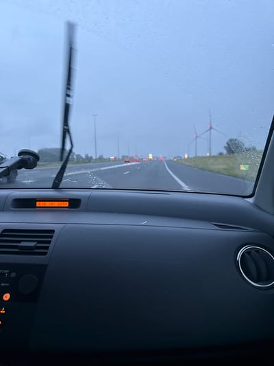The image, taken from inside a vehicle and rotated 90 degrees clockwise, presents a view through a rain-splattered windshield. Raindrops cover the glass, and a windshield wiper is visible at the bottom, suggesting wet weather conditions.
Inside the car, the dark grey dashboard with a textured finish occupies the right side of the frame. A circular air vent with a chrome ring is seen at the top right, and the faint text "SRS AIRBAG" is visible on the dashboard. A vertical orange digital display shows "5:15", "13", and "22.0°", likely indicating the time, an unknown metric, and the temperature. Below this display, part of a car stereo is visible with buttons labeled "TA", "3", "ROM", and "5", and text "CD/CD-R/RW". A suction cup mount for a device is attached to the lower left of the windshield.
Outside, the wet road surface reflects light, and a line of red taillights from distant cars suggests traffic ahead. Several large wind turbines are visible on the horizon, silhouetted against an overcast, light grey sky. Patches of green roadside vegetation are also present. The scene depicts a drive during an overcast late afternoon or early evening in Leidschendam, Netherlands, characterized by rain.