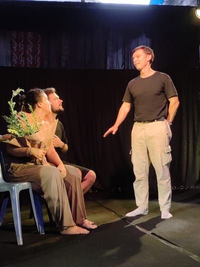 The image captures a scene likely from a performance or rehearsal in Bedulu, Indonesia. Two individuals, a woman and a man, are seated on a light blue plastic chair. The woman is holding a bouquet wrapped in brown paper. The man has his arm around the woman. Both of them gaze to the right. A third man stands to the right, facing the seated pair, with his hand extended in a gesture. He wears a black t-shirt and beige cargo pants with white socks. The backdrop is a dark curtain, with an indistinct decorative element on the left. The flooring appears to be a stage.