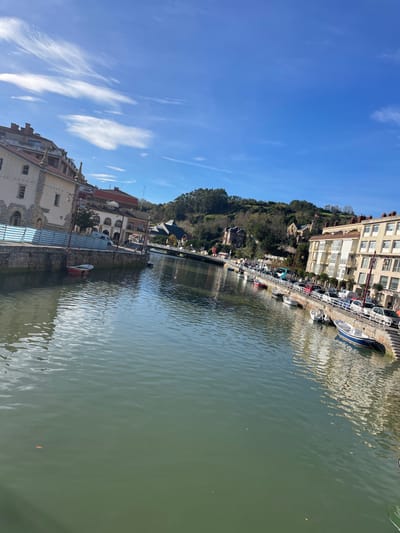 The image depicts a sunlit section of a waterway, identified as a river or estuary in Zumaia, Spain, viewed from a slightly elevated and angled perspective. The sky above is clear blue with scattered, wispy white clouds. The water is a greenish-brown, exhibiting ripples and reflections of the surrounding structures.

On the left bank, a multi-story building with a light-colored facade and a stone foundation stands adjacent to a stone quay. A single red-hulled small boat is moored below this quay. A temporary striped barrier, colored blue and white, is visible along a portion of the quay wall.

The right bank features a stone promenade lining the waterway, along which multiple small boats, including vessels with blue and white hulls, are moored. Behind the promenade, a street contains parked vehicles, specifically cars and a blue bus-like vehicle. Multi-story buildings with balconies are visible beyond the street. A set of stone steps descends from the promenade into the water. Faint human figures are discernible on the promenade.

In the background, a multi-arched bridge spans the waterway. Beyond the urban structures, hills densely covered with green foliage rise, with additional buildings interspersed among the trees.