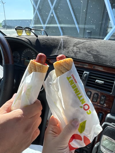 In the foreground of the image, two hands hold hot dogs wrapped in paper. The hot dog on the left is partially visible, showing a red sausage in a bun with some red sauce. The paper wrapper has text that includes "www.royal-petrol.kz". The hot dog on the right is also wrapped in paper with the word "CONTINENT" and "market" printed on it in yellow and red. In the background, the interior of a car is visible, including the dashboard with a speedometer, the steering wheel, air vents, and car controls. A pair of sunglasses with yellow lenses rests on the dashboard. Outside the car, a bright, sunny sky is visible, along with some structures and a large truck. The presence of "royal-petrol.kz" suggests a location related to petrol stations, and the overall context points to a roadside stop, potentially in or near the city of Turkistan, Kazakhstan, a common route for long-distance travel where such amenities are found.