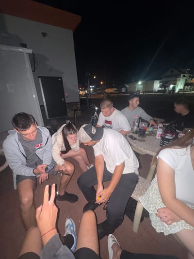 In Ocean City, United States of America, a group of young adults is gathered around an outdoor table at night. The setting appears to be an establishment with a stucco-like exterior and an orange-trimmed roof. The table is set with drinks, including a cup with a dark liquid and a straw, a bottle of water, and a can of Wawa. Several individuals are holding and looking at their mobile phones. One person in the foreground is making a "peace" sign with their fingers, and another is holding a yellow object, possibly a lighter. The individuals are dressed casually in t-shirts, hoodies, shorts, and one person is wearing a floral-patterned skirt. The lighting is dim, consistent with nighttime, and streetlights and building lights are visible in the background. The ground is paved with a reddish-brown material. A bicycle is visible behind the table, and a white van and other vehicles are in the distance.