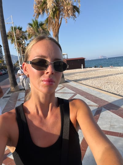 The media file is a vertical, close-up selfie of a young woman with blonde hair and sunglasses, taken outdoors during the day. She is wearing a black tank top and is positioned in the foreground. The background shows a beach with pebbles, the sea, and a clear blue sky, suggesting a sunny day. Palm trees are also visible, along with patterned paving stones and a structure that appears to be part of a building or boardwalk. The sunlight casts strong shadows, indicating it's likely midday or late afternoon. The woman's pose and direct gaze suggest she is taking the photo herself, possibly at a beach resort or promenade in l'Alfàs del Pi, Spain. There is no visible text in the image.