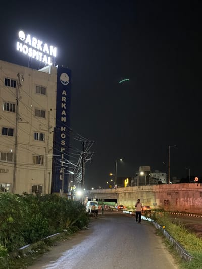 At night, a white building with illuminated "ARKAN HOSPITAL" signage stands on the left side of the image. The building has multiple windows and electrical wiring runs along its facade and in front of it. A vertical blue sign with white lettering, also reading "ARKAN HOSPITAL" and featuring a circular logo, is attached to the building. A person in a light-colored shirt and dark pants walks on a paved road in the foreground, facing away from the camera and towards the right. The road curves to the right and is bordered by a grassy area and a concrete wall. In the background, a highway overpass is visible, with traffic and streetlights. There are also other buildings and a speed limit sign with "40" in red. A faint green curved light trail is visible in the dark sky above. The overall scene depicts a street in Shamshabad, India at night.