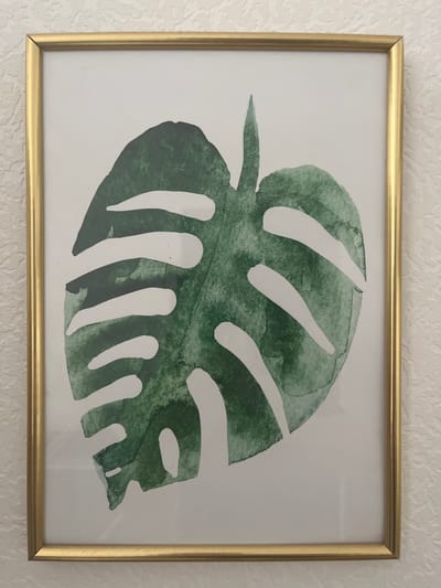 A framed artwork featuring a green monstera leaf is displayed on a textured, off-white wall. The frame is thin and metallic gold. The leaf itself is rendered in watercolor with variations in green tones, and features the characteristic holes of a monstera. The artwork is positioned centrally within the frame. The background wall has a rough, stucco-like texture. The image is a close-up shot, focusing on the artwork and the wall it is mounted on. There are no people or other objects visible in the frame. The location is identified as Bugry, Russia.