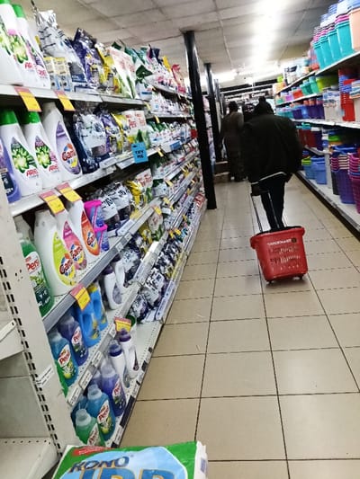 This image captures the interior of a supermarket or grocery store aisle. The perspective is from slightly low, looking down a long, well-stocked aisle.
On the left side of the aisle, numerous shelves are filled with laundry care products. These include liquid detergents in various plastic bottles (white, green, blue, purple) and powdered detergents in bags. Visible brands include "Ariel," "Gama," and "Persil." The packaging features bright colors and bold lettering. Several yellow price tags or promotional labels are attached to the front edge of the shelves. A small blue sign with the text "TRY ME" is visible on one of the lower shelves, suggesting a sample or new product promotion. On the very bottom left, partially cropped, a large bag or package with the text "KONO" is visible on the floor.
On the right side of the aisle, another set of shelves displays an assortment of household plasticware, primarily buckets, bowls, and containers. These items are stacked and arranged, showcasing a variety of bright colors such as blue, pink, purple, red, and green.
In the mid-ground, a person is walking away from the camera, pulling a red shopping basket with wheels and an extendable handle. This person is wearing a dark jacket, possibly black or dark green, with a white stripe on the sleeve, and dark trousers. The red shopping basket clearly displays the brand name "QUICKMART" in white lettering. Further down the aisle, two other individuals are also walking away, less distinctly visible in the background. One appears to be wearing a brown top and patterned bottom.
The floor of the aisle is tiled with light-colored, possibly beige or light grey, square tiles separated by dark grout lines. Overhead, the ceiling appears flat and light-colored, with several overhead lighting fixtures providing illumination. Dark support pillars are strategically placed along the aisle, one prominently standing in the middle section. In the far background, a sign or part of a display mentions "Camster." The store appears to be well-lit and organized, presenting a typical retail environment.