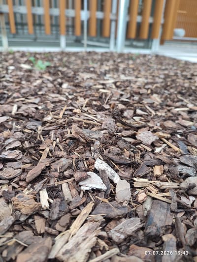 The image is a close-up, low-angle shot of a ground covered with bark mulch. The mulch consists of various shades of brown and gray pieces of wood, some with visible bark texture. In the background, out of focus, is a light-colored fence or barrier made of vertical wooden planks, some appearing orange-brown and others a lighter wood color, separated by white vertical supports. A sliver of green vegetation is visible in the mulch, near the fence. The timestamp "07.07.2026 19:43" is overlaid in the bottom right corner of the image. The scene depicts an outdoor area, possibly a park, garden, or landscaped public space in Immenstadt im Allgäu, Germany. There are no people or animals visible.