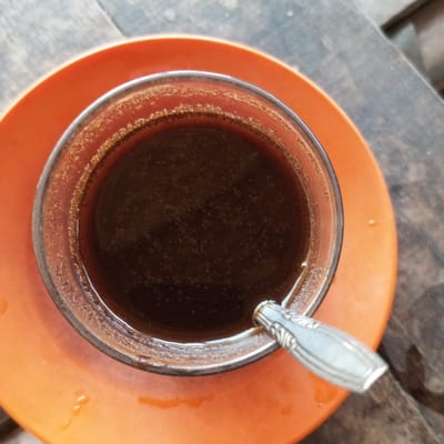An overhead shot shows a glass of dark liquid, likely coffee, set on a bright orange saucer, and a small silver spoon resting in the glass. The setting appears to be an outdoor wooden surface, possibly a table. The location is Matangmeunye, Indonesia.
