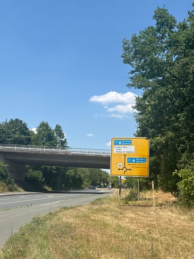 The image shows a yellow directional sign on a bright, sunny day. The sign indicates directions to several German cities and points of interest: "Nürnberg", "Bamberg", "Erlangen-West", "Hafen", "Klinikum Am Europakanal", "Nürnberg Würzburg", and "Neustadt a.d.Aisch Herzogenaurach". The sign also features road numbers "73" and "3", along with directional arrows and a roundabout symbol.
The scene is set outdoors, with lush green trees on the left and dry, tall grass on the right. A concrete bridge or overpass runs diagonally across the middle of the image, casting a shadow on the road below. The sky is a vibrant blue with a few scattered white clouds. The perspective is from the side of the road, looking towards the sign and the bridge. There are some vehicles visible on the road in the distance, and a couple of people are seen on bicycles near the bottom of the image. The overall impression is of a bright, clear day in a semi-rural or suburban area in Germany, possibly near a highway. The presence of road signs with German text and city names confirms the location. The time of day appears to be daytime, likely late morning or afternoon, given the strong sunlight.
