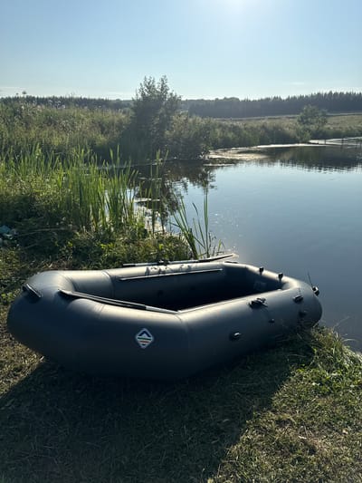 An inflatable boat is parked on the grassy bank of a calm body of water. The boat is dark gray, with oars visible inside. A small, diamond-shaped logo with red, green, and white stripes is visible on the side of the boat. The water reflects the blue sky and the surrounding greenery. Tall reeds and grasses grow along the water's edge. In the background, a dense line of trees stretches across the horizon, with a clear blue sky above. The sun is shining brightly, casting shadows on the grass. The location is the city of Bolshie Shady, Russia. There are no people or animals visible in the image. The scene depicts a tranquil, natural environment.