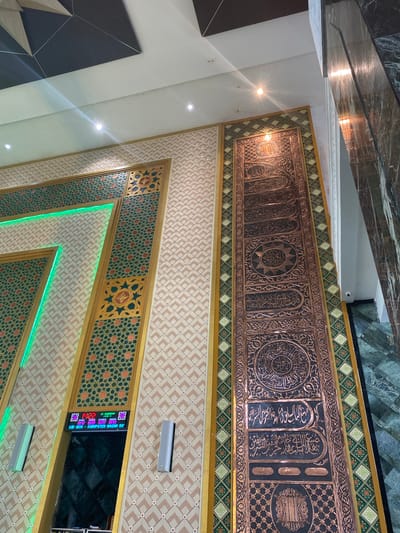 The image depicts the interior of a building, likely a mosque, in Aluekambuk, Indonesia. The walls are adorned with intricate geometric patterns and Islamic calligraphy. A prominent feature is a tall, narrow panel on the right, decorated with ornate metalwork featuring Arabic script. The ceiling has a modern geometric design. There is a digital display on the wall showing prayer times and a date in both Gregorian and Hijri calendars. Two gray speakers are mounted on the wall. The floor appears to be made of polished dark stone with a green marble pattern. No people are visible in the image.