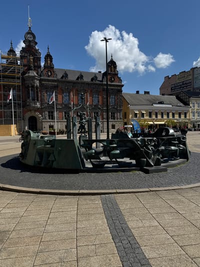 The image shows a bronze sculpture in front of a large brick building in Malmö, Sweden. The sculpture is abstract and features geometric shapes and stylized figures. The building behind it has ornate details, including statues, a clock tower, and flags. Scaffolding is visible on the building's left side. A paved area with cobblestone accents surrounds the sculpture, and other buildings are visible in the background. A clear blue sky with scattered white clouds is overhead. Text on a building in the background refers to businesses such as "J. ALTHEN'S" and "GRAND RESTAURANT HIPPODROME." This location is likely in a central part of Malmö, possibly near the Stortorget (Main Square) or in a historical area.