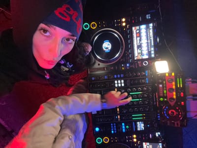 A single individual is depicted operating professional DJ equipment in a dimly lit, colorfully illuminated indoor setting, specifically in Redlands, United States of America. The subject, a person with dark hair, dark eyes, and fair skin, wears a dark hooded garment, a navy blue beanie with red text, and a light-colored outer layer partially visible on their left arm. Their left hand is positioned on a multi-channel Pioneer DJM mixer, actively adjusting a control knob, indicated by the green level indicators. The individual's gaze is directed towards the camera. The DJ setup includes at least two Pioneer CDJ digital media players and the central mixer, with various illuminated screens and controls indicating active operation. A circular jog wheel on the left CDJ displays "66 BPM" and "PLAY." A separate red and black effect unit, possibly a Pioneer RMX-1000, with a small white auxiliary light source attached, is visible on the far right. Ambient lighting casts strong red, pink, and blue hues across the scene and the subject's face. The floor beneath the subject appears to be a textured red surface.