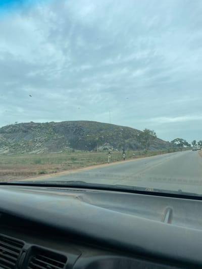 A rocky outcrop rises beside a highway near Kujama, Nigeria. Two people stand by the roadside, one wearing a yellow vest. A few trees grow on the hill. A vehicle is driving on the highway. The sky is overcast. The photo was taken from inside a vehicle, looking through the windshield.