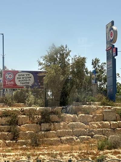 The image depicts a street-level view of a commercial area under a clear blue sky. In the foreground, a dry, overgrown embankment of rough-hewn stone blocks is visible. A dark metal fence with vertical bars runs along the bottom of the image. Dominating the midground is a large billboard sign for "Ten" with a circular logo featuring a smiling red face. The billboard, which is in Hebrew, translates to "How do you like your RUSTO?" and advertises coffee. To the right of the billboard, a tall, blue signpost with multiple panels displays pricing information and logos, typical of a gas station. The "Ten" logo is also prominently featured on this signpost. Behind the billboard and signpost, various trees and bushes create a green backdrop. In the far background, a utility pole with a dark arm can be seen on the left. The overall impression is of a commercial establishment in a somewhat arid, sunny environment. The presence of the "Ten" branding, common in gas stations and convenience stores in Israel, suggests a location along a major road. Research indicates that "Ten" operates numerous stations throughout Israel, including in Dimona, a city in the Southern District of Israel.