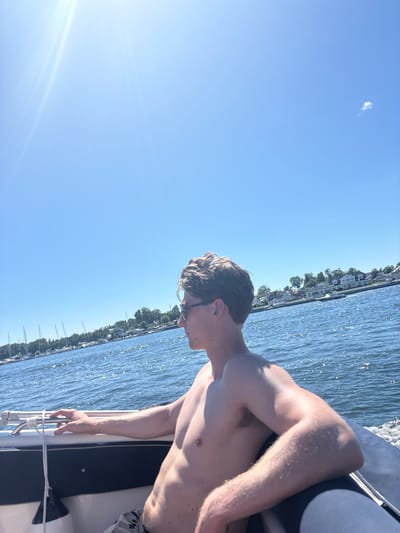 An outdoor, medium shot shows a shirtless young Caucasian male from the waist up, wearing sunglasses and black shorts, sitting on the edge of a boat. He is facing to the left of the frame, looking out at the water. The boat is moving, as evidenced by the wake behind it and the spray of water on the right side of the frame. The water is a deep blue, with sunlight glinting off the surface. In the background, a shoreline is visible, lined with houses and trees. Several sailboats are docked in the distance. The sky is a clear, bright blue with a few wispy clouds. The image is taken on a sunny day. This scene is set in Tønsberg, Norway.