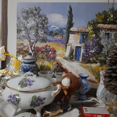 A still life arrangement is presented in front of a painting. The painting depicts a sunlit rural scene with a white house, blue doors and shutters, trees, and a lavender field. In the foreground, there is a white porcelain serving dish with a lid, decorated with purple floral motifs and gold trim. Beside it sits a small, brown, teddy bear wearing a gold ribbon. Various decorative objects, including glass bowls, a pinecone, and a blue and white ceramic pot, are also part of the arrangement. The image appears to be taken indoors, possibly in a residential setting. There is no indication of the city of Vila Verde, Portugal.