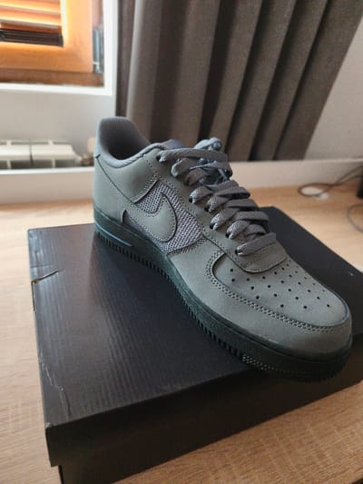A single dark grey athletic shoe, resembling a Nike Air Force 1 model, is presented at an angle, resting on a dark, rectangular cardboard shoebox. The shoe features a combination of suede-like or nubuck material on the toe box, eyelets, and heel, with a textured mesh material visible on the midfoot panel beneath the matching dark grey Nike swoosh. Dark grey laces are tied on the shoe, and the toe box contains multiple perforations. The sole is a solid dark grey, almost black, with visible traction patterns. The shoebox displays minor surface abrasions and indentations. The objects are positioned on a light-colored wooden surface with a visible grain. In the background to the left, a light brown wooden window frame with horizontal slats and a white radiator is visible. To the right, a dark brown or grey draped curtain partially covers a wall, with a faint black cable visible on the floor. The scene is located in the city of Ribnovo, Bulgaria.
