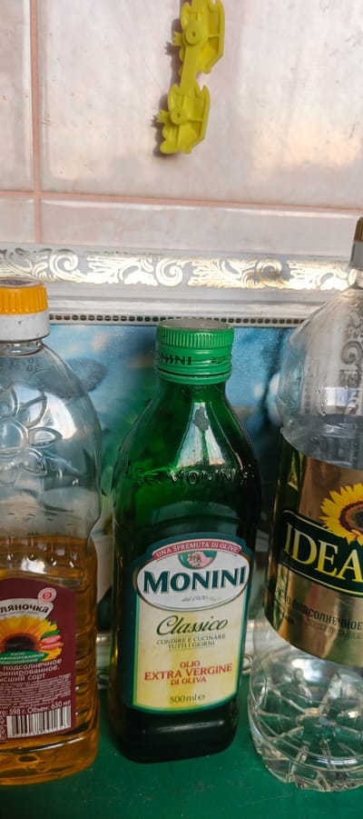 A close-up, vertical, eye-level shot shows three plastic bottles and a yellow plastic toy duck on a tiled wall in Tikhvin, Russia. The bottle on the left, partially filled with golden oil, has a yellow cap and a label with a sunflower. The bottle in the center, filled with dark green liquid, has a green cap and a label with "MONINI CLASSICO OLIO EXTRA VERGINE DI OLIVA." The bottle on the right, also partially filled with clear liquid, has a partially visible label with a sunflower. The toy duck is suspended from the wall. The background is a tiled wall with a decorative border.