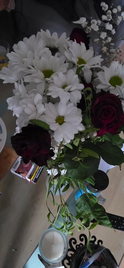 A bouquet of flowers, primarily white daisies with green centers, is arranged in a vase. Two dark red roses are interspersed among the daisies. Some delicate white baby's breath flowers are also visible. The bouquet is positioned on a light-colored, reflective surface, possibly a table. To the left of the bouquet, a pack of cigarettes is partially visible. The background is dark and out of focus. In the lower portion of the image, a white candle in a glass holder and a dark decorative object are present. The scene appears to be indoors. The specific location in the city of Bergen, Germany, cannot be determined from the provided image.