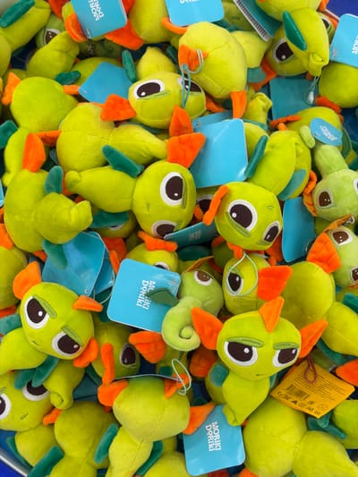 The image shows a close-up view of a pile of plush toys. These toys are light green with orange beaks and green wings. Each toy has a white and brown embroidered eye. Many of the toys have a blue rectangular tag attached, which reads "MORIKI DORIKI." A yellow tag is also visible near the top right of the frame, displaying product information and a barcode. The toys are densely packed together, filling the frame. The image suggests the toys are being sold in a store or market. There is no other information available to verify the location or context.