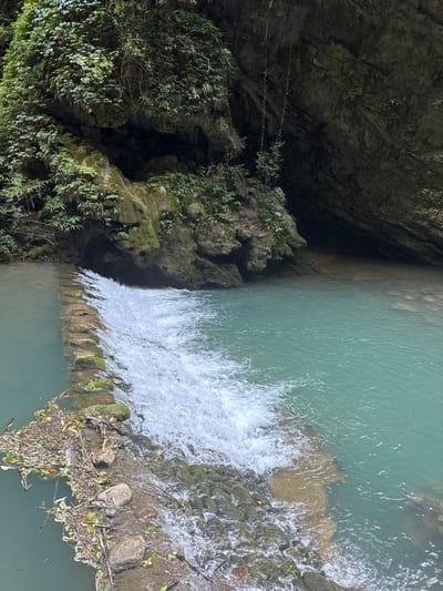 The image depicts a serene natural scene within the Sanguansi, China, area. A small waterfall cascades over a man-made stone barrier, creating white, foamy water. The water is a vibrant turquoise color, and the surrounding rocks are covered in lush green moss and vegetation. Cliffs, partially obscured by dense foliage and hanging vines, rise on either side of the water. Debris, including leaves and twigs, has accumulated on the left side of the stone barrier, indicating the natural flow of water and organic matter. The overall impression is one of untouched natural beauty and tranquility.