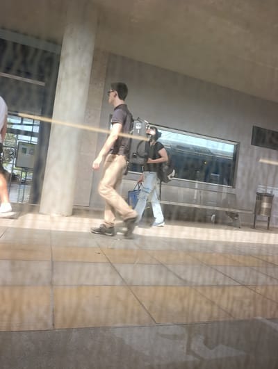 An exterior shot shows two individuals walking on a tiled platform in Palmela, Portugal. The individual on the left, viewed from the side, is wearing a dark short-sleeved shirt and light brown pants, carrying a black backpack. They are also wearing dark athletic shoes and glasses. The second individual, also viewed from the side, is wearing a dark short-sleeved shirt, light blue distressed jeans, and dark sneakers, and is carrying a blue bag. They have dark hair and appear to be wearing a face mask. The background features a concrete pillar, a glass partition reflecting a train and a trash receptacle. The platform is illuminated by sunlight, casting shadows.