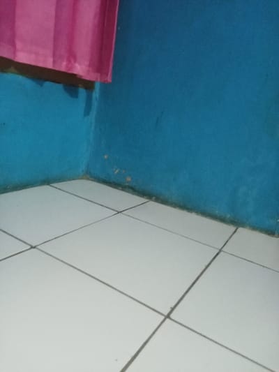This image depicts an interior corner of a room in Ciputri, Indonesia. The walls are painted a solid medium blue/teal color. A section of a pink fabric curtain is visible hanging in the upper left quadrant, casting a shadow on the wall below it. The floor is composed of light-colored, possibly white, square ceramic tiles with dark grout lines forming a grid pattern. Along the base of the walls, where they meet the floor in the corner, there is a visible accumulation of dark discoloration or dirt. Additionally, several small, irregular yellowish-orange spots are present on the blue wall, primarily concentrated near the corner and closer to the floor. No people or specific actions are depicted.