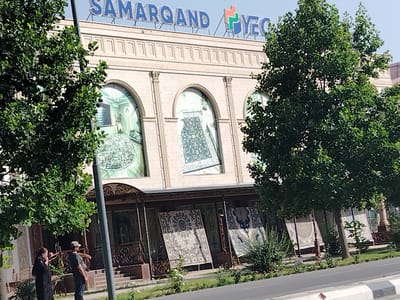 The image shows the "Samarqand" store in Shahrisabz City, Uzbekistan. The building has a pale beige facade with arched windows displaying large rugs. Above the windows, the store's name is written in blue lettering. The entrance to the store is at ground level, with decorative metal railings and several large rugs displayed on stands. Two people are visible near the entrance: a woman in a black outfit talking on a cell phone and a man wearing a cap and a dark shirt, pointing towards the building. Lush green trees partially obscure the building from view, and a road runs in front of it. The sky is clear and blue.