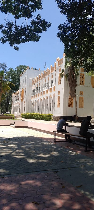 The scene depicts an outdoor daytime environment in Tudor ward, Kenya, under a clear blue sky. A multi-story building with white walls and distinctive yellow-orange decorative elements, consistent with Swahili architectural style, dominates the background. The building features arched ground-level recesses, upper-story rectangular windows (several open), a crenelated parapet, and multiple visible air conditioning units. Green foliage from trees, including palm trees, frames the upper portion of the image and flanks the building. In the mid-ground, a paved area displays intricate tree shadows, transitioning from a reddish-brown pattern with interlocking circles to a darker grey interlocking brick pattern. A low, light pink barrier wall separates the paved area from the building's foundation, which is lined with green hedges. Two male individuals are seated on a wooden bench with dark metal legs in the mid-foreground, facing away from the camera. The individual on the left wears a light-colored shirt, and the individual on the right wears a dark-colored shirt; both are looking downwards. No direct interaction between them is discernible.