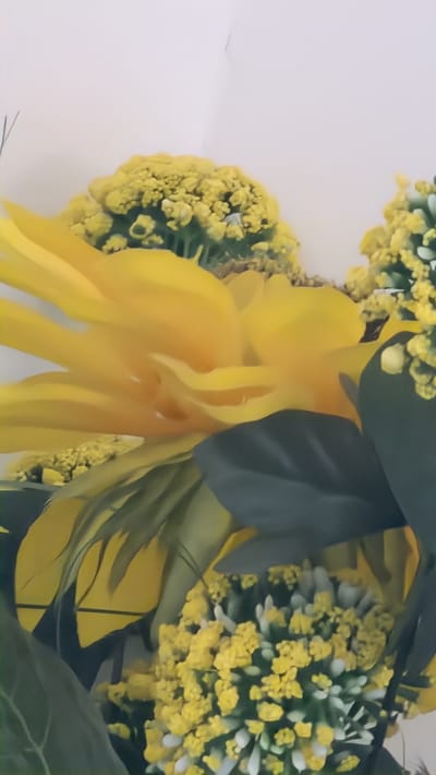 The image shows a close-up of a yellow floral arrangement against a white background. The arrangement includes a large, multi-petaled yellow flower, possibly a sunflower or dahlia, surrounded by smaller clusters of yellow and white flowers, and dark green leaves. The composition is dense, filling the frame. The location in the city of Lincoln, United Kingdom is not discernible from the image. There are no people, subjects, actions, or interactions visible. The only objects present are the floral arrangement and the background.